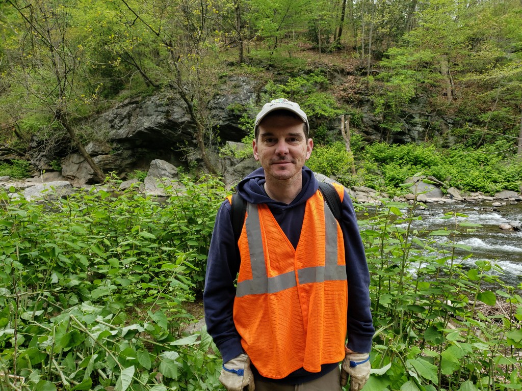 A man in an orange safety vest and ballcap stands with a backdrop of various bushes, vines and trees, with a creek flowing between rocks.