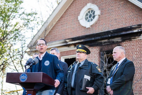 Gov. Josh Shapiro stands at a lectern outside a brick building with a uniformed officer and several other people standing nearby.